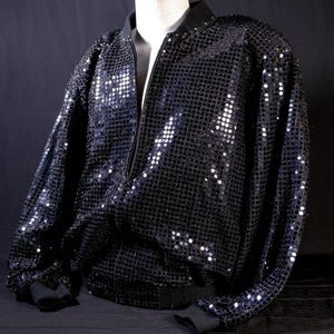 Può includere: Giacca bomber nera con paillettes e chiusura a zip completa. La giacca ha polsini e orlo a coste neri. Le paillettes sono fitte, creando un effetto scintillante. La giacca è esposta su un manichino.