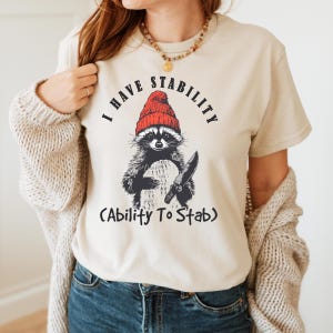 May include: Beige t-shirt featuring a raccoon wearing a red beanie and holding a knife. The text above the raccoon reads "I HAVE STABILITY" and below it, "(Ability To Stab)". The raccoon is black and white.