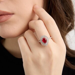 May include: A silver ring featuring a pear-shaped, deep red gemstone, surrounded by a halo of smaller, clear stones. The ring is on a person's finger, with the hand in focus. The background is blurred.