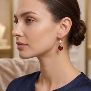 Puede incluir: Un pendiente colgante de oro con una pequeña perla blanca en la parte superior y una gran piedra preciosa de color rojo intenso, tallada en forma de pera y facetada, suspendida debajo. El pendiente se sujeta con un gancho de oro pulido.