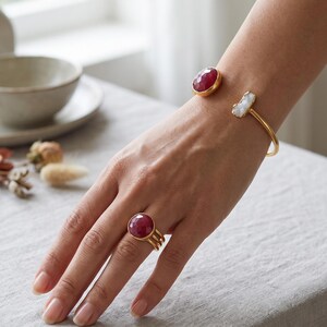 May include: A gold-toned open cuff bracelet featuring a faceted round deep red stone and a rectangular white stone, paired with a matching gold-toned ring featuring a faceted round deep red stone set on a triple-band shank design.