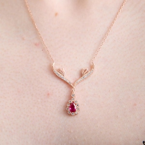 Puede incluir: Collar de oro rosa con colgante. El colgante presenta una piedra preciosa roja en forma de lágrima, rodeada de pequeños cristales transparentes. El collar tiene una cadena delicada y un diseño decorativo sobre el colgante.