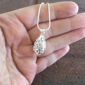 May include: A silver filigree pendant necklace with a teardrop-shaped cage design. The necklace is hanging from a silver chain.