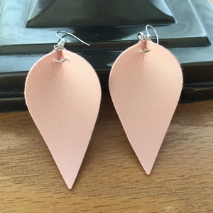 May include: A pair of light pink teardrop-shaped earrings with silver-colored ear wires. The earrings have a textured surface and are displayed on a wooden surface.