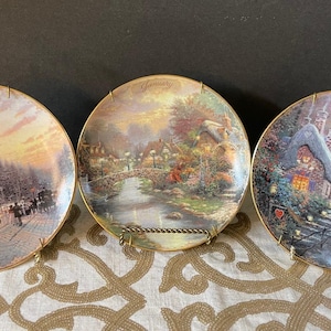 May include: Three decorative plates with gold rims, each depicting a different winter scene. The plates are labeled "December", "January", and "February". Each plate is displayed on a gold-colored stand.