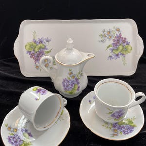 May include: A white porcelain tea set with a floral design. The set includes a rectangular tray, a small teapot with a lid, two teacups, and two saucers. The floral design features purple and white flowers with green leaves, and gold trim.