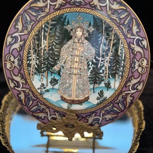 May include: A decorative plate featuring a woman in traditional attire set against a winter forest scene. The plate has a purple and gold border with ornate designs. The woman is wearing a detailed coat and headdress. The plate is displayed on a decorative metal stand.
