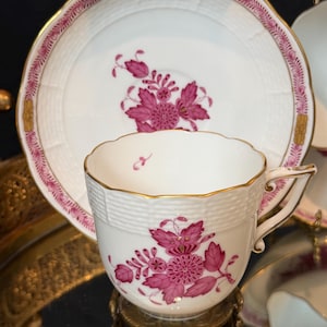 May include: White teacup and saucer set with a pink floral design. The cup and saucer have gold trim and a textured basketweave pattern. The floral design is centered on the cup and saucer.