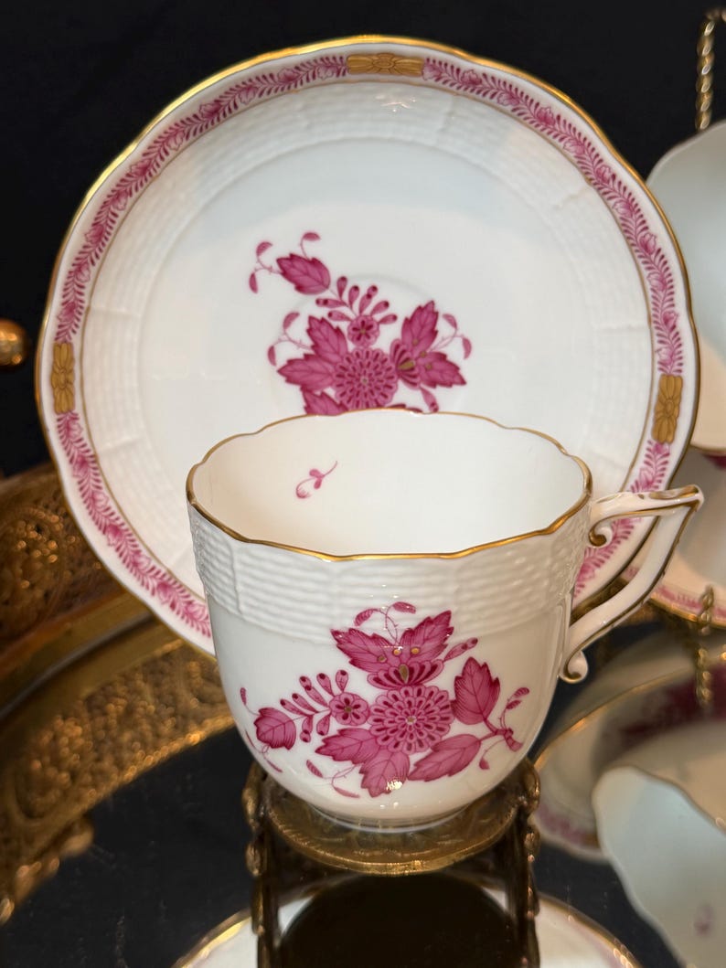 May include: White teacup and saucer set with a pink floral design. The cup and saucer have gold trim and a textured basketweave pattern. The floral design is centered on the cup and saucer.