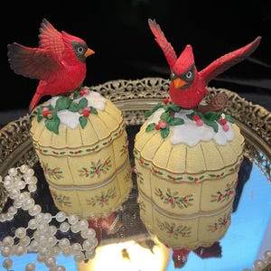 May include: Two decorative trinket boxes with red cardinal bird figurines perched on top. The boxes are yellow with a floral design and are sitting on a mirrored tray. A strand of pearls is in the foreground.