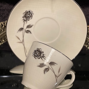 May include: Cream-colored teacup and saucer set with a black rose design. The cup and saucer have a textured surface and a dark trim. The rose design is on both the cup and the saucer. The set is displayed on a reflective surface.