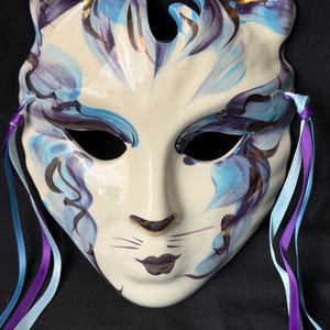Peut inclure: Un masque en céramique en forme de chat, avec une base crème, orné de motifs floraux bleus et violets et d'accents dorés. Le masque comporte des trous pour les yeux, un nez et une bouche, avec des rubans bleus et violets attachés.