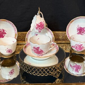 May include: A set of vintage teacups and saucers. The white porcelain cups and saucers feature a pink floral design and gold trim. The teacups are displayed on a mirrored surface and a decorative gold stand, creating an elegant presentation.