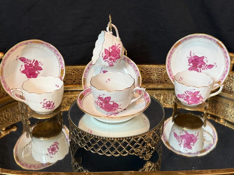 May include: A set of vintage teacups and saucers. The white porcelain cups and saucers feature a pink floral design and gold trim. The teacups are displayed on a mirrored surface and a decorative gold stand, creating an elegant presentation.