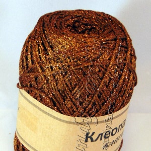 May include: A ball of brown yarn with gold metallic threads. The yarn is wrapped with a tan paper label with text in a foreign language. The yarn is tightly wound and has a textured appearance.