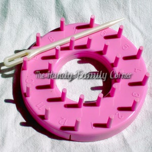 May include: A pink circular knitting loom with evenly spaced pegs and a white plastic yarn needle. The loom has numbers around the edge and the text "The Handy Family Corner".