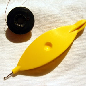 Könnte beinhalten: Eine gelbe Schiffchenspitze mit einem gebogenen Haken und einer schwarzen Fadenspule. Das Schiffchen hat eine Tropfenform mit einem kleinen Loch und einem spitzen Ende. Die Fadenspule ist schwarz, mit etwas Faden sichtbar.