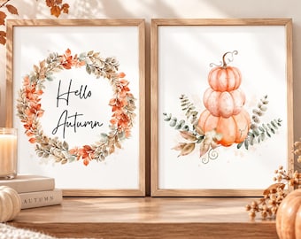 May include: Two framed wall art prints with light wood frames. The left print features a wreath of orange and green autumn leaves surrounding the text Hello Autumn. The right print displays a stack of three pale orange pumpkins with foliage.