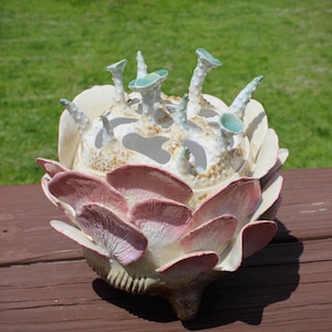 May include: A ceramic sculpture featuring a cluster of pink, fan-shaped mushroom gills surrounding a central white vessel. The top of the vessel is adorned with several small, upright, light green, textured stalks with flat, circular tops.