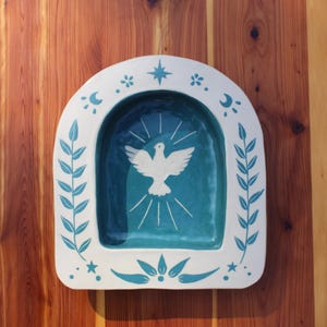 Puede incluir: Una hornacina de pared de cerámica blanca con interior color cerceta que presenta un gráfico de paloma blanca con líneas radiantes. El marco arqueado está decorado con ramas de hojas, estrellas, lunas crecientes y motivos florales.