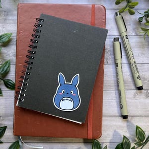 Puede incluir: Un cuaderno de espiral negro con una pegatina de un personaje de dibujos animados azul, colocado sobre un cuaderno marr&oacute;n. Dos bol&iacute;grafos de punta fina negros etiquetados Micron 03 y Micron 05 est&aacute;n situados a la derecha.