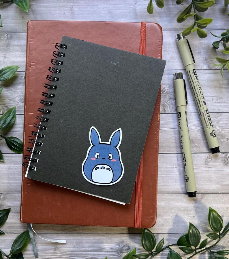 Puede incluir: Un cuaderno de espiral negro con una pegatina de un personaje de dibujos animados azul, colocado sobre un cuaderno marr&oacute;n. Dos bol&iacute;grafos de punta fina negros etiquetados Micron 03 y Micron 05 est&aacute;n situados a la derecha.