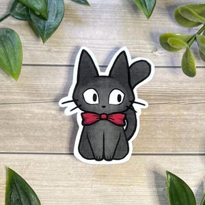 Puede incluir: Una pegatina troquelada que presenta una ilustraci&oacute;n estilizada de un gato peque&ntilde;o de color gris oscuro con grandes ojos blancos y una pajarita roja brillante. El gato aparece sentado con la cola curvada hacia arriba y un borde blanco.