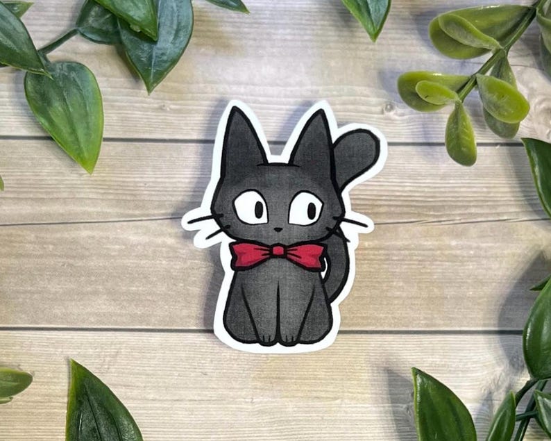 Puede incluir: Una pegatina troquelada que presenta una ilustraci&oacute;n estilizada de un gato peque&ntilde;o de color gris oscuro con grandes ojos blancos y una pajarita roja brillante. El gato aparece sentado con la cola curvada hacia arriba y un borde blanco.