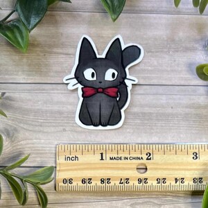 Puede incluir: Una pegatina troquelada de un gato negro con grandes ojos blancos y una pajarita roja. El gato est&aacute; colocado sobre una regla de madera, mostrando que la pegatina mide unos 6,3 cm de alto y ancho, con un borde blanco alrededor del dibujo.