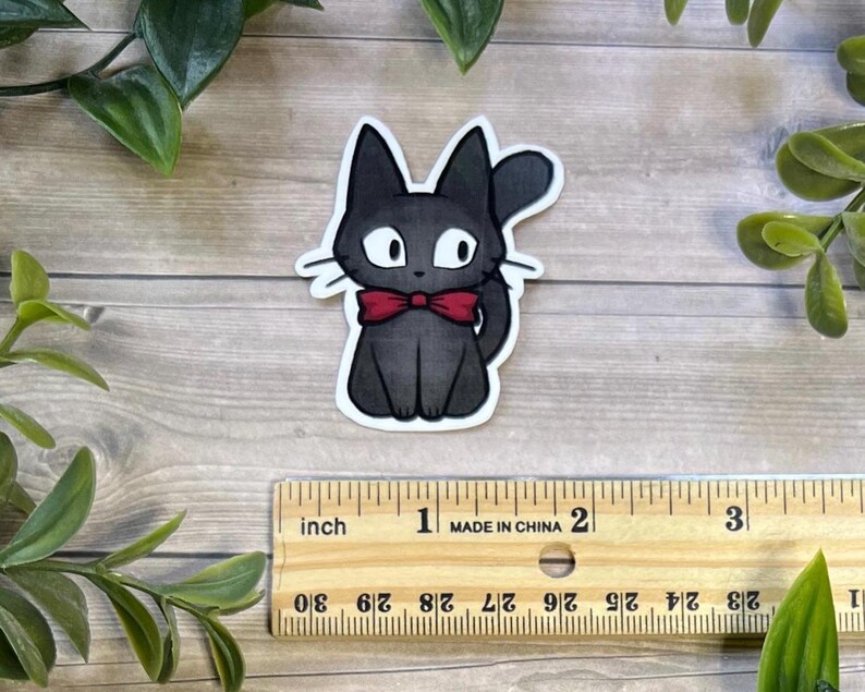 Puede incluir: Una pegatina troquelada de un gato negro con grandes ojos blancos y una pajarita roja. El gato est&aacute; colocado sobre una regla de madera, mostrando que la pegatina mide unos 6,3 cm de alto y ancho, con un borde blanco alrededor del dibujo.