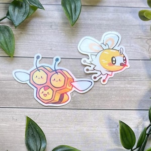 May include: Two die-cut vinyl stickers featuring cartoon characters. One sticker depicts a yellow and orange honeycomb-shaped creature with three smiling faces and antennae. The second sticker shows a small yellow and white bee-like character.