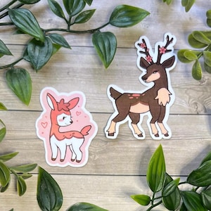 May include: Two die-cut stickers featuring deer illustrations. One sticker shows a small, light pink and white fawn with heart accents. The second sticker depicts a larger, brown deer with dark antlers adorned with small pink cherry blossom flowers.