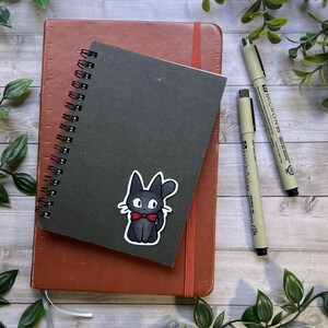 Puede incluir: Un cuaderno de espiral negro con una pegatina de un gato negro con pajarita roja, colocado sobre un cuaderno marr&oacute;n m&aacute;s grande. Dos bol&iacute;grafos Micron 05 de color beige con tapas negras est&aacute;n situados a la derecha sobre una superficie de madera.
