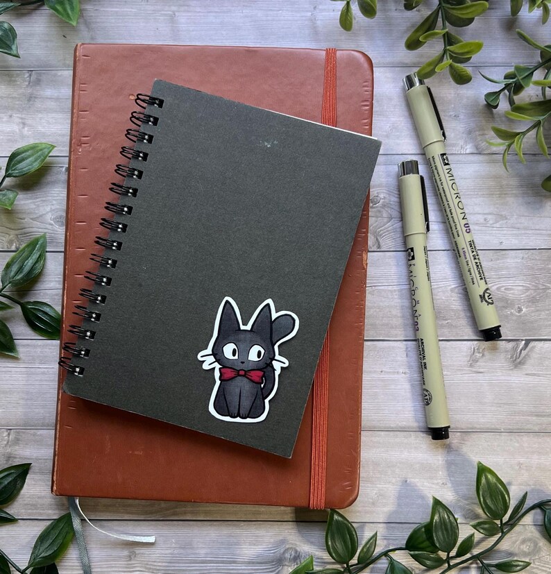 Puede incluir: Un cuaderno de espiral negro con una pegatina de un gato negro con pajarita roja, colocado sobre un cuaderno marr&oacute;n m&aacute;s grande. Dos bol&iacute;grafos Micron 05 de color beige con tapas negras est&aacute;n situados a la derecha sobre una superficie de madera.