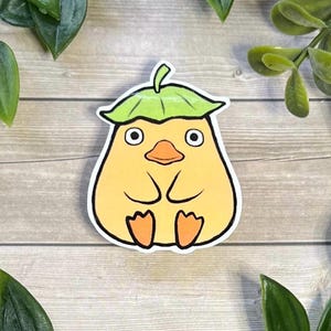 Puede incluir: Una pegatina troquelada que muestra una criatura redonda y amarilla parecida a un pato, con un pico ancho naranja y pies naranjas. La criatura tiene ojos grandes y circulares y lleva una hoja verde como sombrero, con un borde blanco grueso.