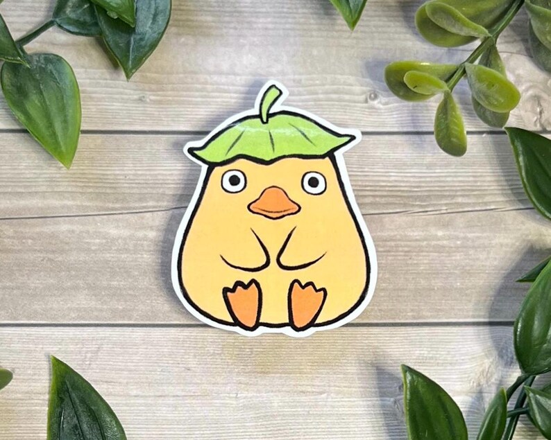 Puede incluir: Una pegatina troquelada que muestra una criatura redonda y amarilla parecida a un pato, con un pico ancho naranja y pies naranjas. La criatura tiene ojos grandes y circulares y lleva una hoja verde como sombrero, con un borde blanco grueso.