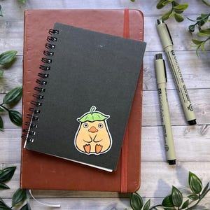 Puede incluir: Un cuaderno de espiral negro con una pegatina de una criatura redonda de color naranja claro con un sombrero de hoja verde, colocado sobre un cuaderno marr&oacute;n m&aacute;s grande con dos bol&iacute;grafos negros de punta fina a la derecha.