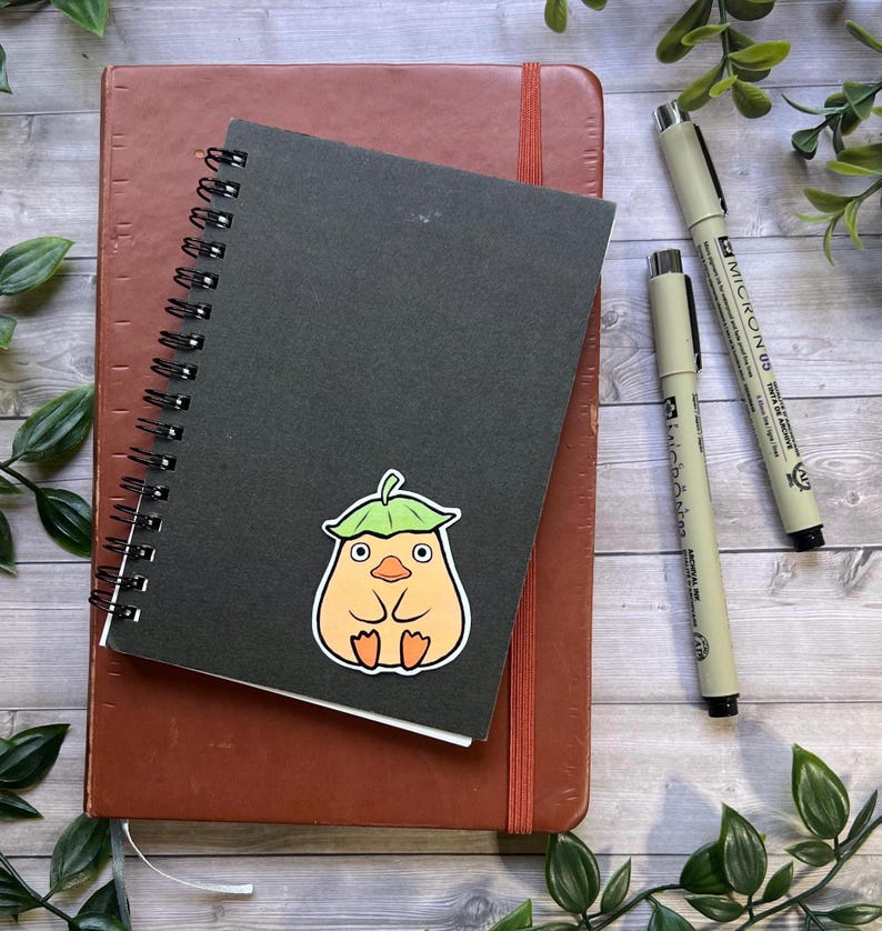 Puede incluir: Un cuaderno de espiral negro con una pegatina de una criatura redonda de color naranja claro con un sombrero de hoja verde, colocado sobre un cuaderno marr&oacute;n m&aacute;s grande con dos bol&iacute;grafos negros de punta fina a la derecha.