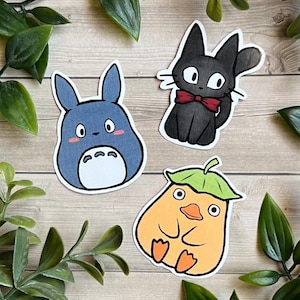 Puede incluir: Tres pegatinas troqueladas con personajes de dibujos animados: una criatura azul con marcas blancas en el vientre, un gato negro con pajarita roja y una figura naranja similar a un pato con un sombrero de hoja verde sobre madera clara.