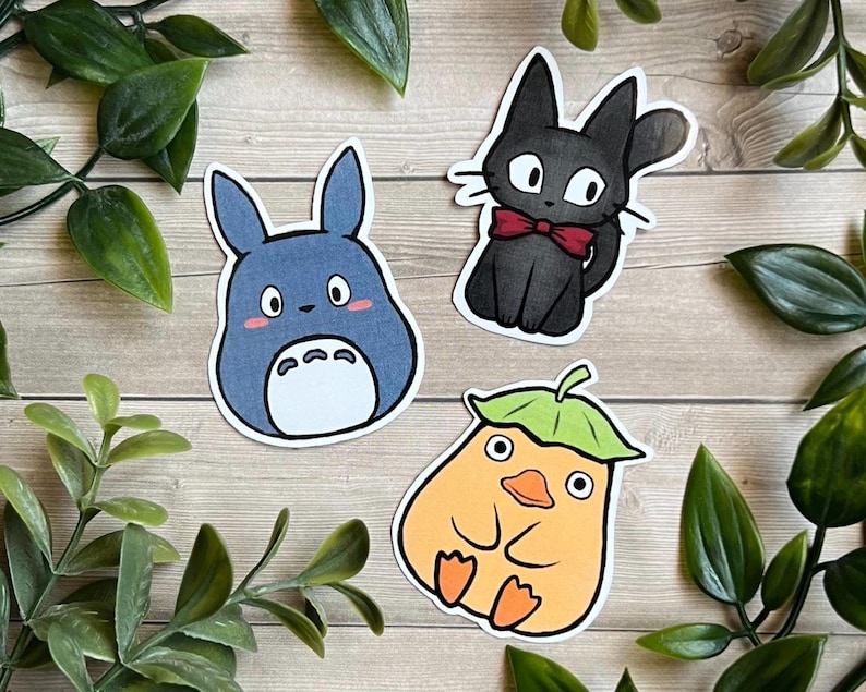 Puede incluir: Tres pegatinas troqueladas con personajes de dibujos animados: una criatura azul con marcas blancas en el vientre, un gato negro con pajarita roja y una figura naranja similar a un pato con un sombrero de hoja verde sobre madera clara.
