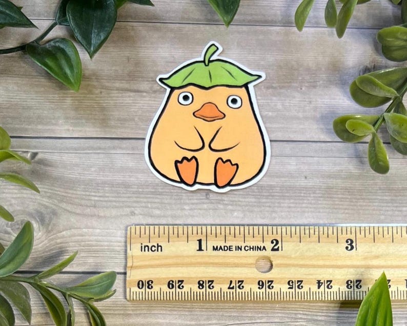 Puede incluir: Una pegatina troquelada que muestra un personaje de dibujos animados con un cuerpo de color naranja claro en forma de pera, un sombrero de hoja verde y pies naranjas, colocada sobre una regla de madera que mide unos 5,7 cm de ancho.