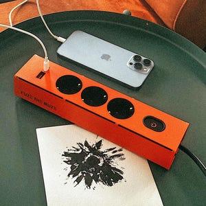 以下が含まれることがあります： 「FUZE THE MUZE」と書かれたオレンジ色の電源タップが、3つのコンセントと電源ボタンを備え、濃い緑色の表面に置かれています。シルバーのスマートフォンが接続されています。黒い花の模様が描かれた白いカードが隣にあります。