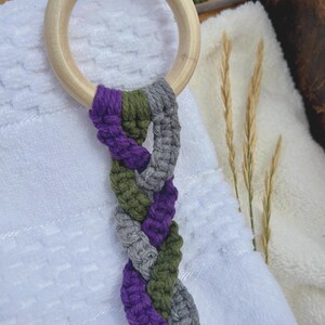 May include: A smooth, light-colored wooden ring attached to a braided cord made of thick yarn in alternating shades of deep purple, olive green, and heather gray. The braided textile hangs vertically, showcasing a textured, knotted fiber construction.