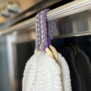 May include: A white kitchen towel hanging from a wooden ring, which is attached to a woven fabric loop featuring a repeating pattern of purple, light green, and grey threads. The loop is draped over a horizontal stainless steel metal bar handle.