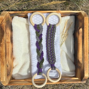 May include: Two macrame towel holders featuring light-colored wooden rings at each end. One holder displays a braided pattern, while the other features a flat knotted design. Both are crafted with purple, olive green, and gray cord fibers.