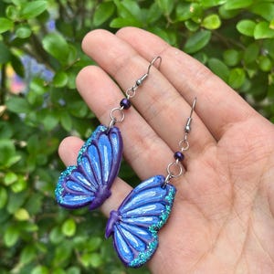 Könnte beinhalten: Ein Paar Ohrhänger mit Schmetterlingsflügel-Anhängern in Violett- und Blautönen. Die Flügel haben eine glänzende Oberfläche mit türkisfarbenen Glitzerakzenten an den Rändern. Jeder Ohrring hängt an einem silberfarbenen Haken mit einer kleinen violetten Perle.