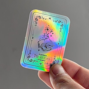 Könnte beinhalten: Ein rechteckiger holografischer Aufkleber mit schwarzem Umriss. Der Aufkleber zeigt den Schriftzug "The Reader" am unteren Rand. Im Inneren befindet sich eine Illustration eines offenen Buches, einer Blume und Sterne. Der Aufkleber hat einen regenbogenfarbenen, schillernden Effekt.