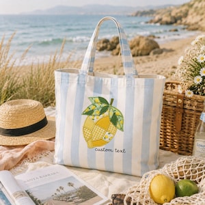 Könnte beinhalten: Eine gestreifte Tragetasche mit blau-weißen vertikalen Streifen. Die Tasche hat einen Zitronenaufnäher und den Text "custom text". Ein Strohhut, ein Magazin und Zitronen liegen am Strand.