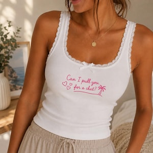 Op de afbeelding: Een wit geribbelde tanktop met kanten afwerking langs de halslijn en schouderbandjes, voorzien van een klein strikje in het midden. De voorkant toont roze tekst Can I pull you for a chat? met drie kleine hartjes en een palmboom.