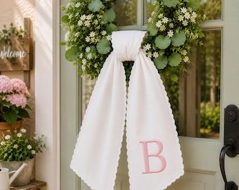 Peut inclure: Une couronne verte circulaire ornée de petites fleurs blanches et de feuilles rondes, agrémentée d'une longue écharpe en tissu blanc. L'écharpe présente des bords festonnés et une grande lettre B brodée en rose sur l'une de ses extrémités.